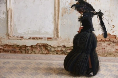 Model wearing a black sculptural pleated skirt and black feather accessory standing against a weathered wall with exposed brick.