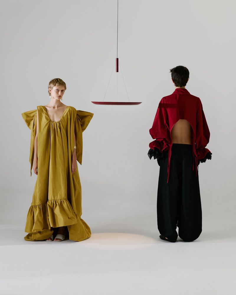 Two models standing against a white seamless background with a hanging red pendant light fixture between them.