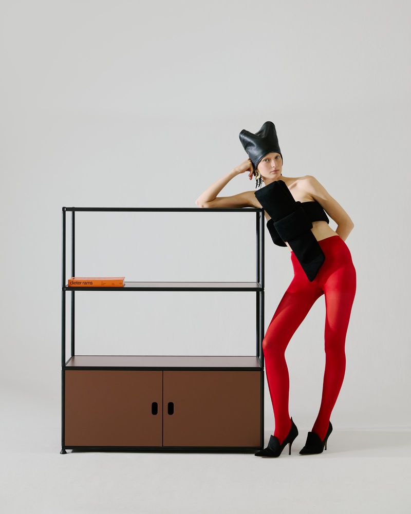 Model Anna Pavlova poses in bright red tights and a sculptural black top beside a minimalist black storage unit on a white background.