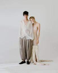 Two fashion models posing together in draped neutral and sheer garments against a white studio backdrop.
