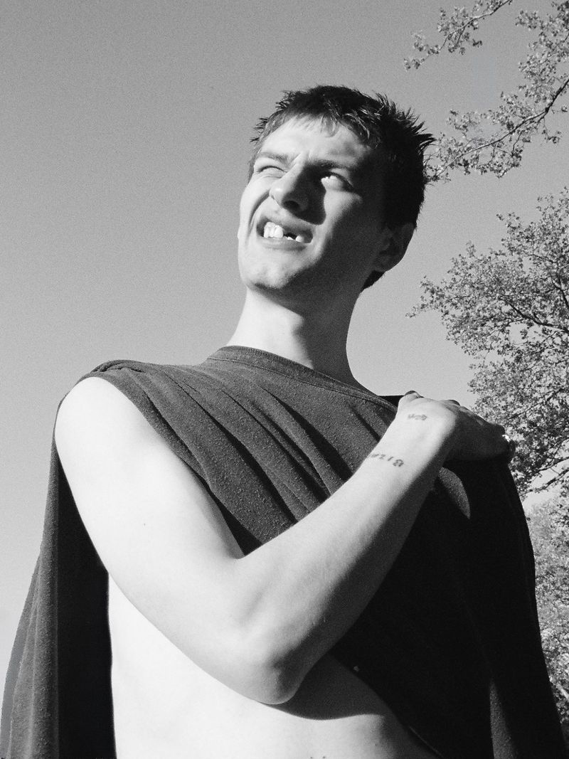 Black and white low-angle portrait of a man squinting upward in draped dark clothing against an open sky with tree branches.