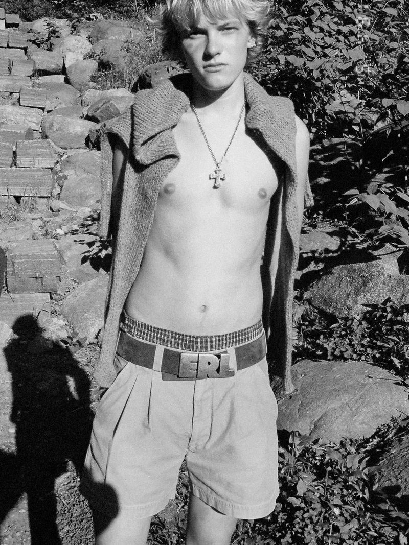 Black and white portrait of a shirtless young blond man wearing shorts, an ERL belt buckle, and a sweater draped over his shoulders.