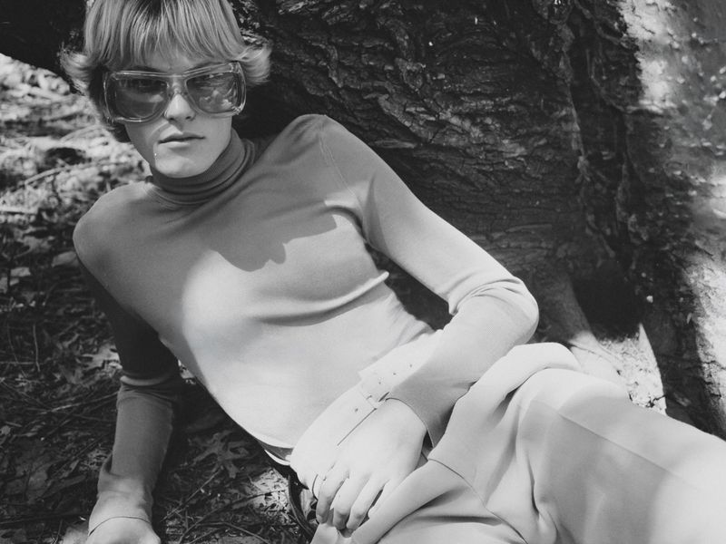 Model wearing oversized sunglasses and a fitted turtleneck reclining against a large tree trunk in black and white.