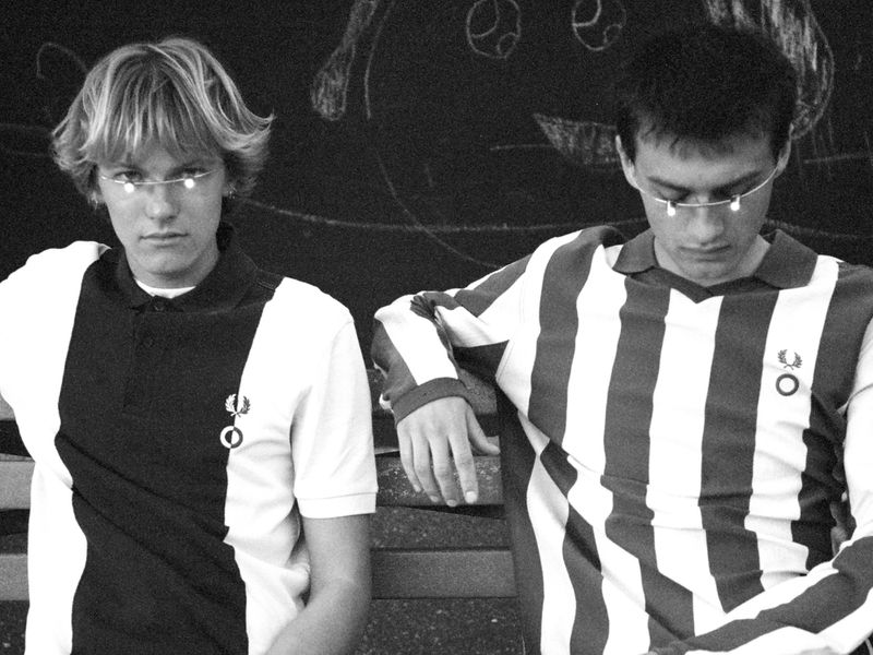 Two male models in striped and color-blocked collared shirts with minimalist wire eyewear seated side by side in black and white.
