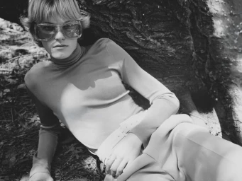 Model wearing oversized sunglasses and a fitted turtleneck reclining against a large tree trunk in black and white.
