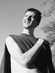 Black and white low-angle portrait of a man squinting upward in draped dark clothing against an open sky with tree branches.