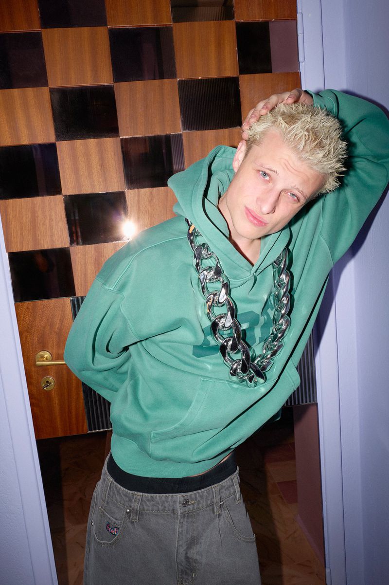 Young man with bleached blonde hair wearing a green hoodie and oversized silver chain necklace leaning against a checkered wood door.