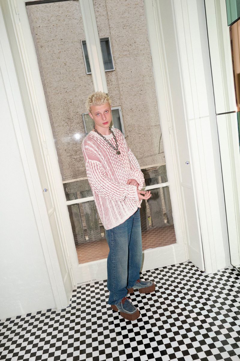 Model wearing an oversized pink textured knit sweater and baggy denim jeans standing on a black and white checkered floor.