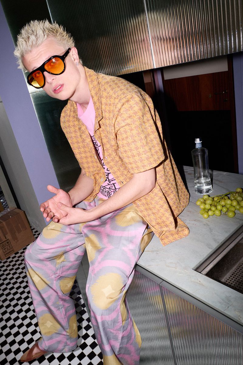 Model with bleached hair and tinted sunglasses sitting on a counter in a patterned shirt and wide-leg trousers.