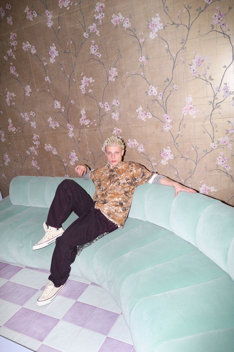 Young man with blonde hair seated on a pale turquoise curved sofa against a floral wallpaper background for Vanity Teen Magazine.