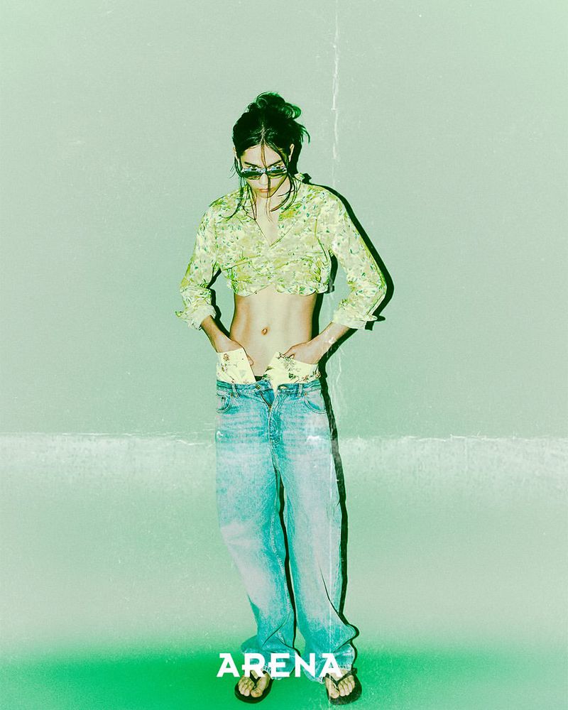 Model Ilias Dosumbaev wearing a cropped floral shirt, sunglasses, and baggy low-rise jeans against a pale green background.