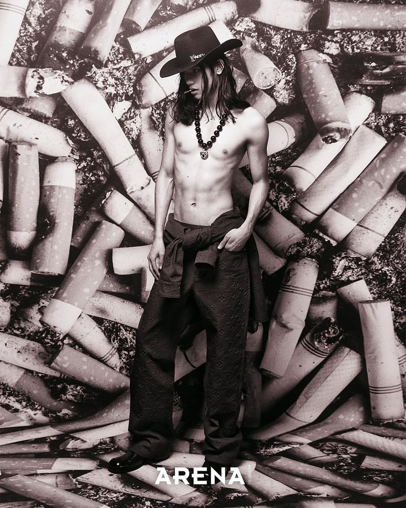 Shirtless model Ilias Dosumbaev wearing a black cowboy hat and trousers against a backdrop of cigarette butts for Arena Homme+.