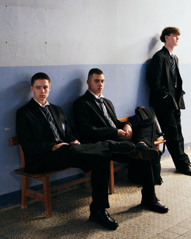 Three young men in black school uniform blazers and striped ties seated on a wooden bench against a blue and grey wall.