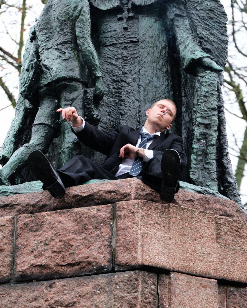 Young man in a black suit and tie smoking a cigarette while reclining on the stone base of a bronze monument.