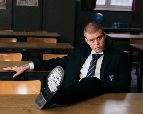 Young man with a buzz cut wearing a dark school uniform blazer and tie resting his foot on a wooden classroom desk.