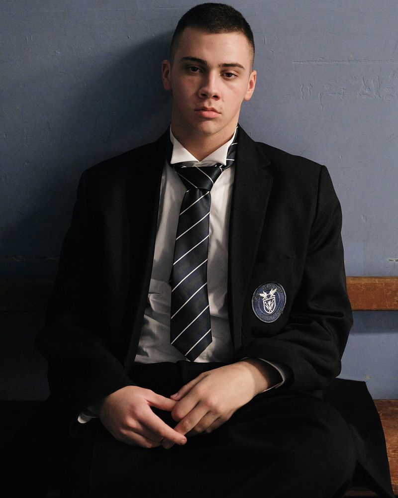 Young man with short cropped hair wearing a black uniform blazer, striped tie, and white collared shirt seated against a blue wall.