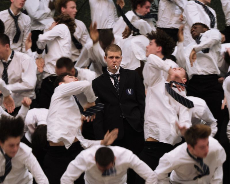 Man in black suit and tie with cigarette standing still amid a dynamic group of men in white shirts