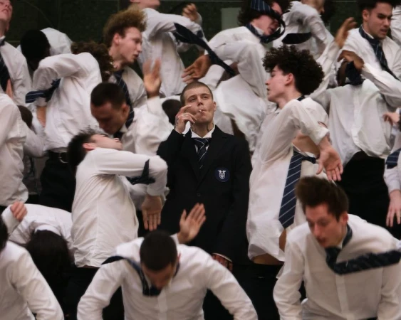 A calm young man in a black school blazer smoking a cigarette stands centered amidst a frantic crowd in white shirts and neckties.