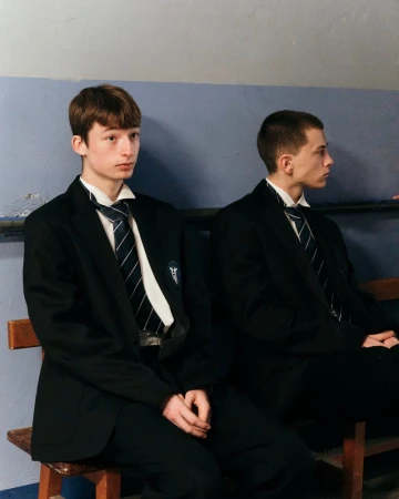 Two young men in black school blazers, white collared shirts, and striped ties seated on a wooden bench against a blue wall.