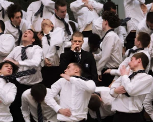 Group of young men in white shirts and ties posing chaotically around a central man in a black blazer smoking.