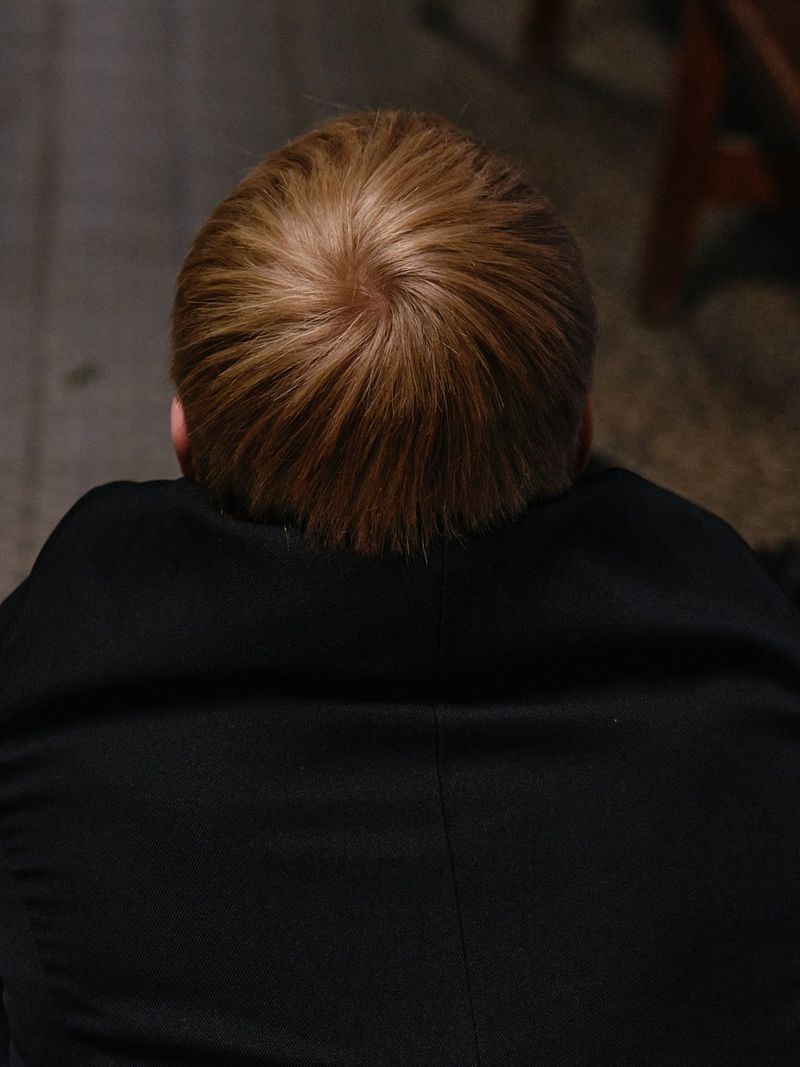 Overhead view of the crown of a person's head with short blond hair wearing a black jacket against a dark floor.