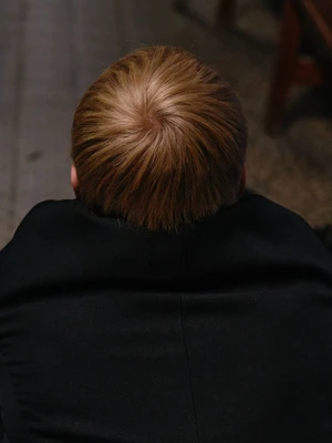 Overhead view of the crown of a person's head with short blond hair wearing a black jacket against a dark floor.