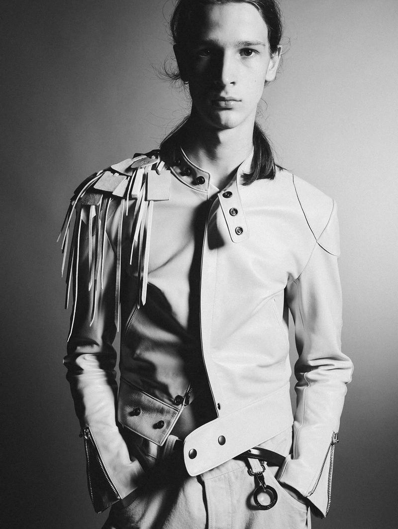 Black and white portrait of a young man wearing a light leather jacket with shoulder fringe detailing against a studio backdrop.