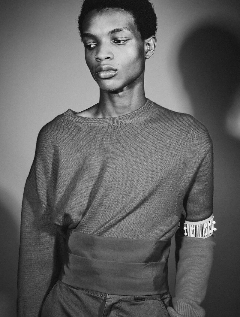 Black and white portrait of a male model in a boatneck knit sweater with an embellished armband against a studio backdrop.