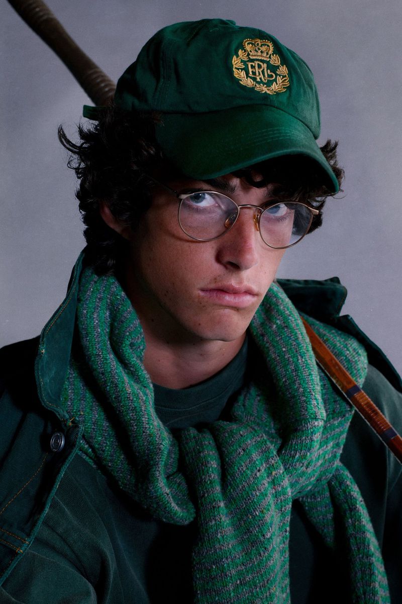 Young man wearing a green embroidered ERL cap, wireframe glasses, and green knit sweater tied over his shoulders.
