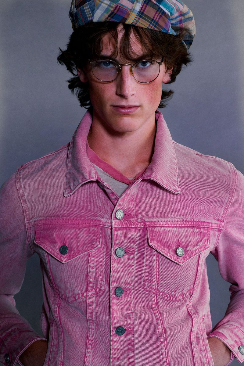Male model wearing wire-frame glasses, a plaid beret, and a faded pink denim jacket against a neutral grey studio background.