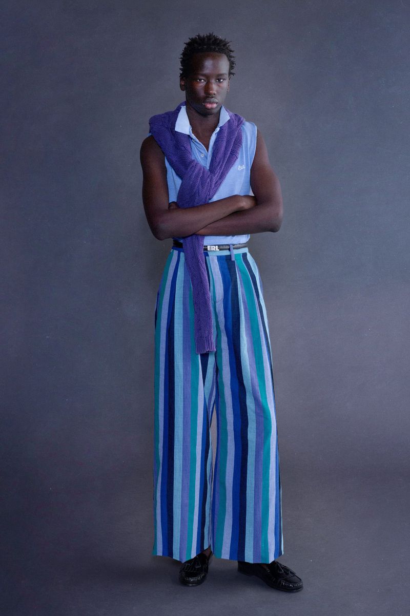 Model wearing wide-leg striped blue trousers with a draped purple knit sweater and light blue top against a gray studio background.