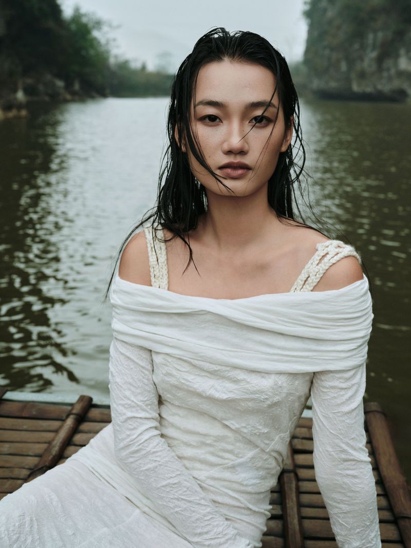 Portrait of an adult woman wearing a white dress while sitting on a bamboo raft in a river with trees and cliffs in the background.