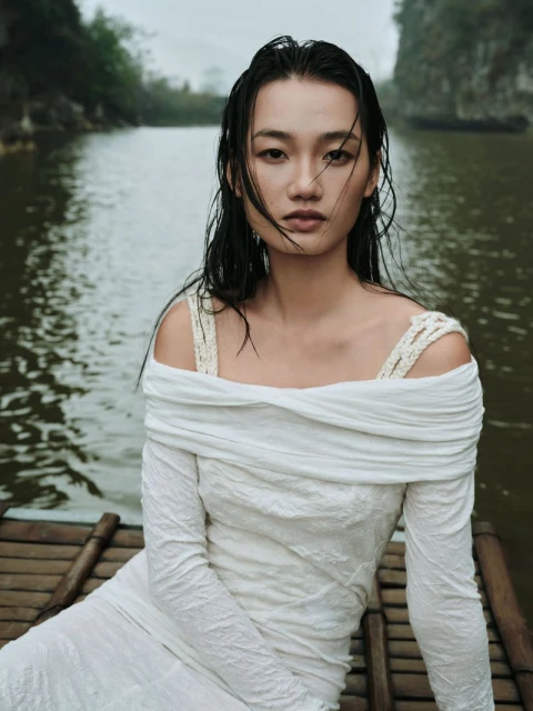 Portrait of an adult woman wearing a white dress while sitting on a bamboo raft in a river with trees and cliffs in the background.