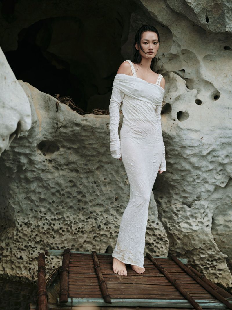 Woman in white dress poses on a wooden walkway in front of a rock cave with textured walls, bathed in natural light, creating an editorial fashion shot.