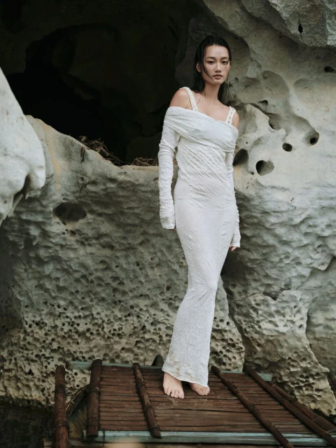 Woman in white dress poses on a wooden walkway in front of a rock cave with textured walls, bathed in natural light, creating an editorial fashion shot.