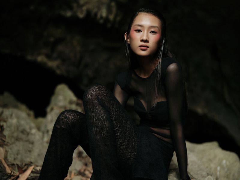 Asian woman in sheer black top and pants poses in front of a dark rocky background with artistic lighting on her face and body.