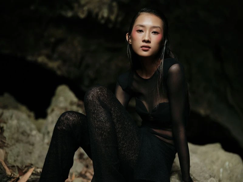 Asian woman in sheer black top and pants poses in front of a dark rocky background with artistic lighting on her face and body.