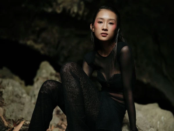 Asian woman in sheer black top and pants poses in front of a dark rocky background with artistic lighting on her face and body.