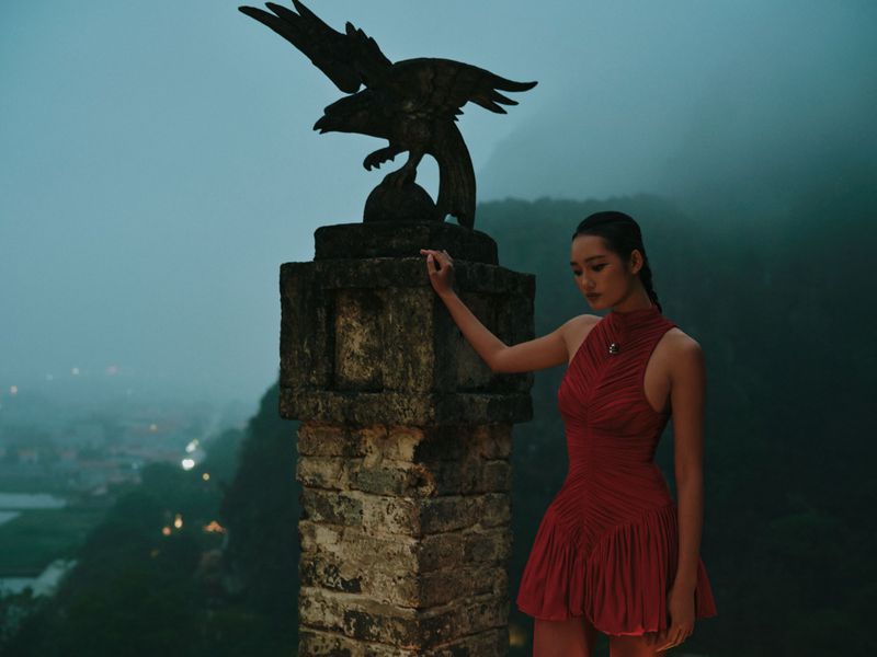 A woman in a red dress poses near a stone pillar topped with an eagle statue, set against a misty mountain backdrop at twilight.