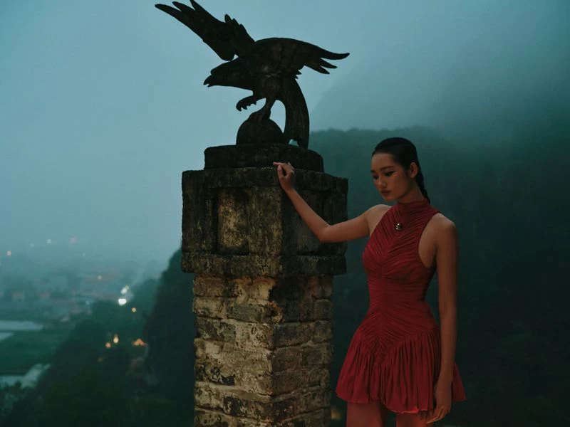 A woman in a red dress poses near a stone pillar topped with an eagle statue, set against a misty mountain backdrop at twilight.