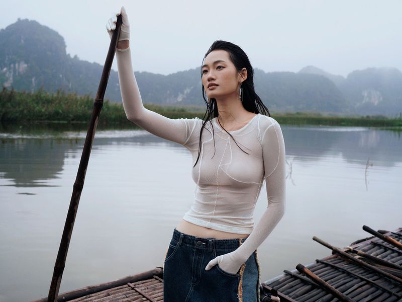 Woman in sheer top and skirt paddles boat on river, mountain landscape, overcast sky, creating a tranquil and atmospheric scene.