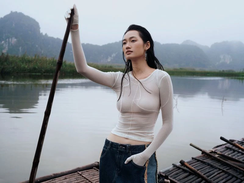 Woman in sheer top and skirt paddles boat on river, mountain landscape, overcast sky, creating a tranquil and atmospheric scene.