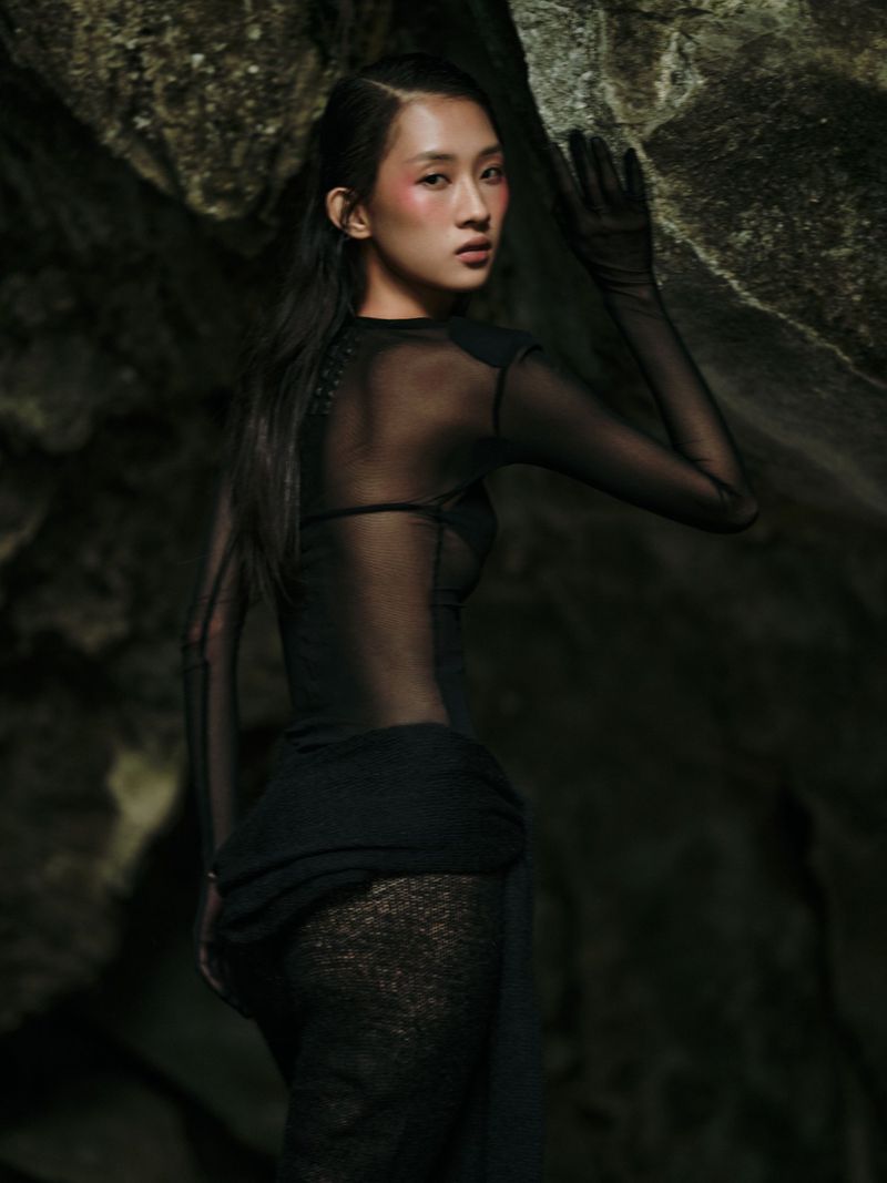 Portrait of an Asian woman in a sheer black dress posing against a rocky background, with visible blush and sleek, dark hair.