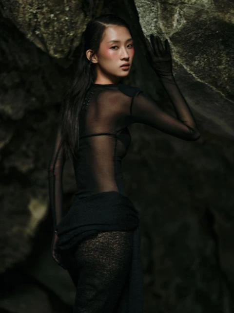 Portrait of an Asian woman in a sheer black dress posing against a rocky background, with visible blush and sleek, dark hair.