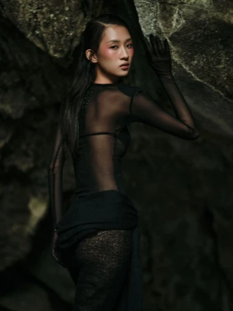 Portrait of an Asian woman in a sheer black dress posing against a rocky background, with visible blush and sleek, dark hair.