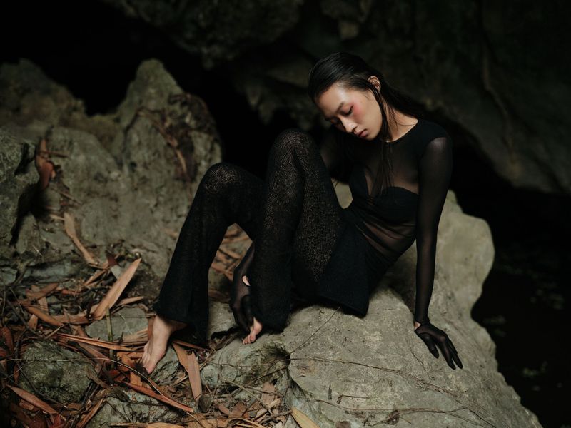 Asian woman in black sheer top and pants poses on gray rock formation with fallen leaves near dark water in dimly lit outdoor scene.