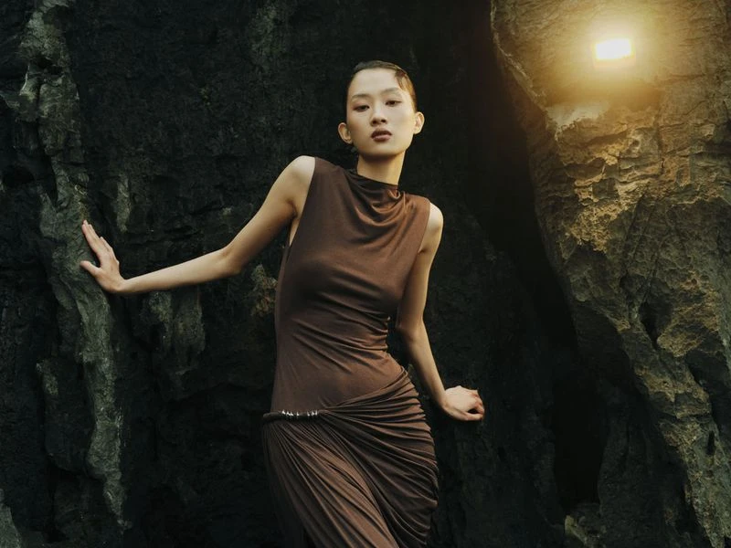 Woman poses in flowing brown dress against a rocky cliff face with a warm, diffused light adding to the natural, earthy tones.