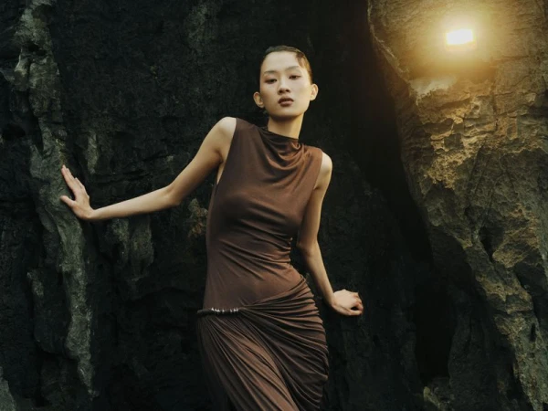 Woman poses in flowing brown dress against a rocky cliff face with a warm, diffused light adding to the natural, earthy tones.