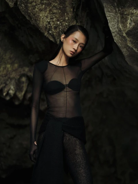 Fashion shot of Asian woman wearing sheer black top and dark skirt posing against rock wall in an indoor environment with dark lighting.