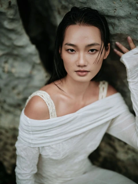 Woman with wet hair poses in a white dress against a grey rock formation, in a natural outdoor setting, looking at the camera.